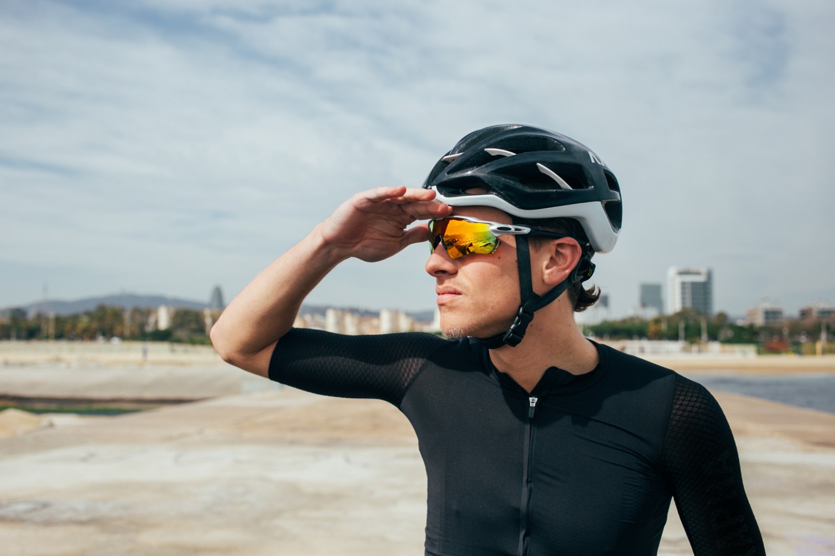 Professional cyclist in all black looks into the distance wearing yellow lenses in glasses with city out of focus in the background
